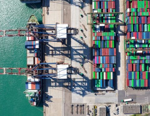 Vertical top down view of cargo containers piled on the quayside and cranes unloading the goods from a ship