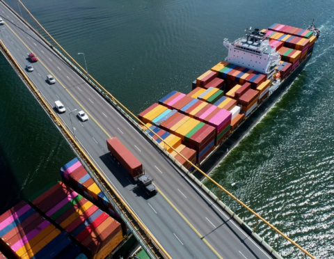 Aerial view of a container ship passing beneath a bridge and semi truck.